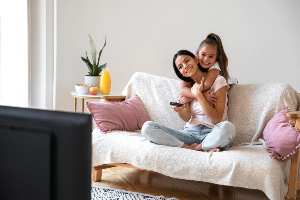 Wer muss Rundfunkgebühren bezahlen? Eine Mutter und ihre Tochter sitzen auf einer Couch und sehen fern.