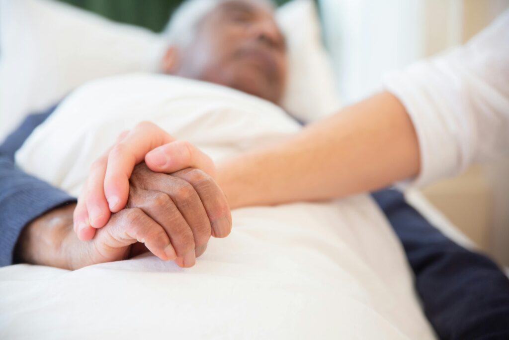 Close-up of two hands clasped together on a bed, conveying caregiving and comfort.
