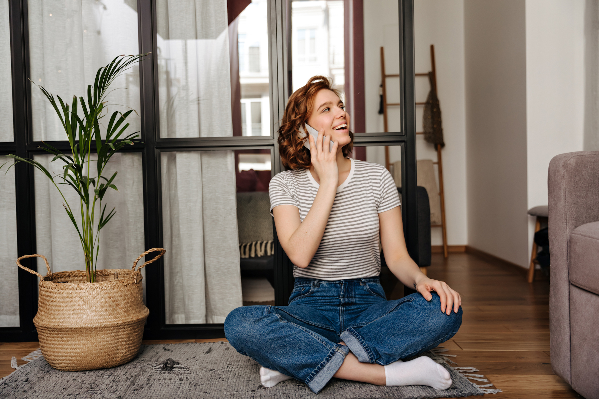 Lovely woman in jeans is sitting on floor in living room and talking on phone