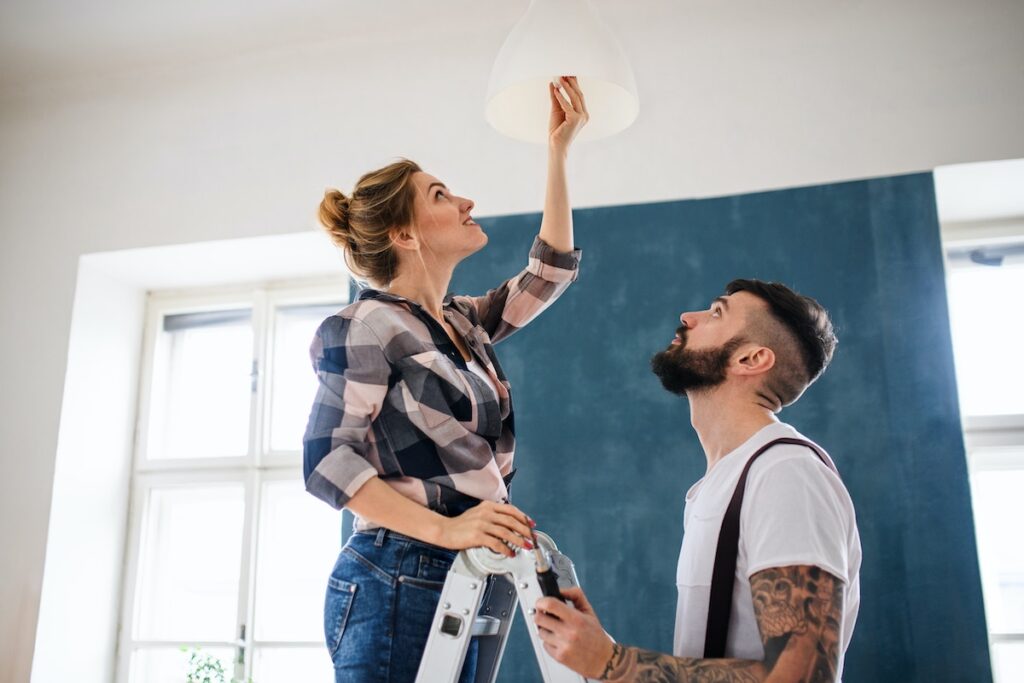 Young couple taking advice for making furnished rentals more eco-friendly: a woman standing on a ladder changing an LED lightbulb, a man holding ladder.