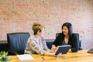 Two women are sitting at a table, talking. One woman is holding a tablet.