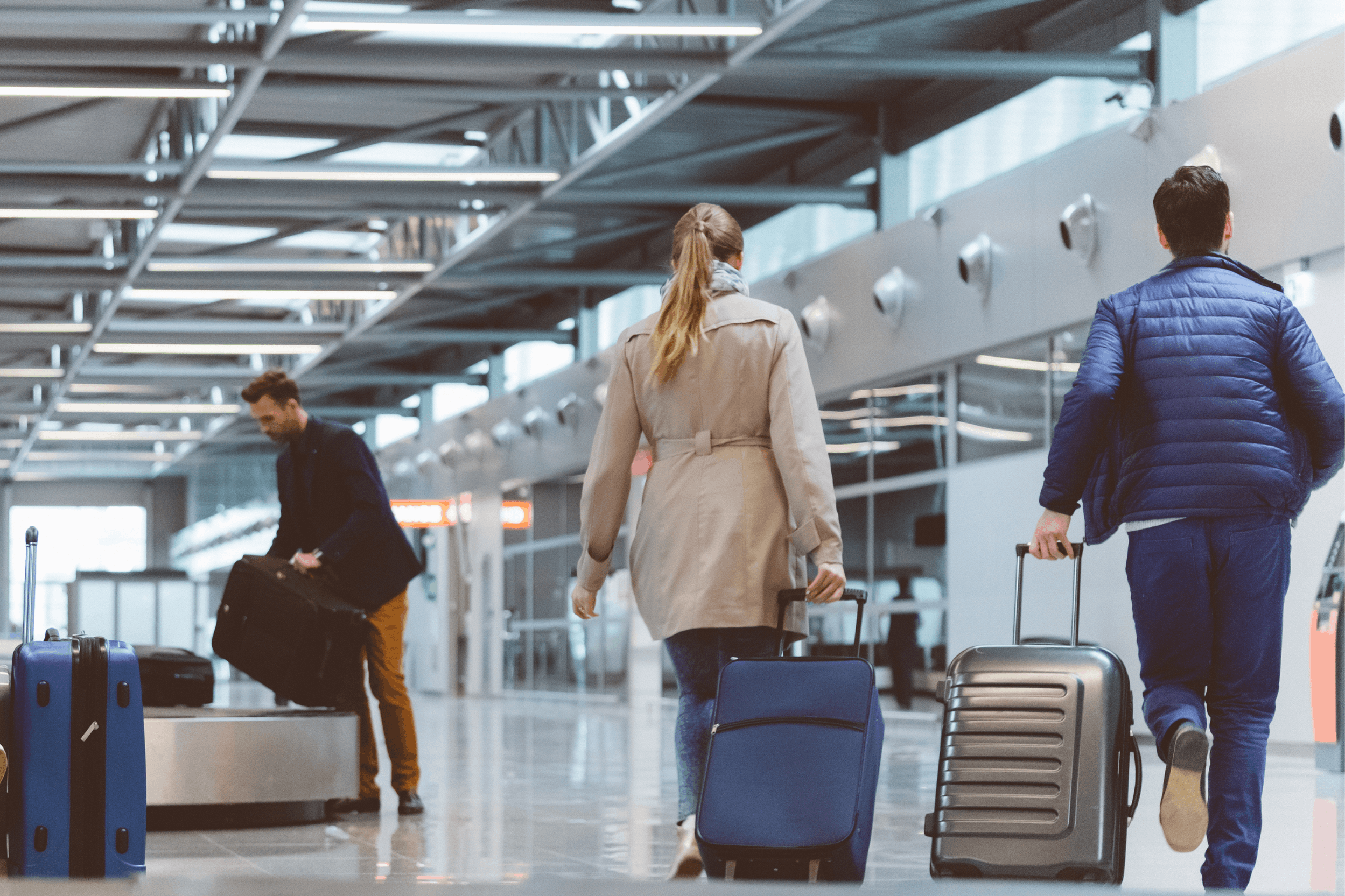 Three travelers with rolling suitcases walk through a bright, modern airport terminal viewed from behind.