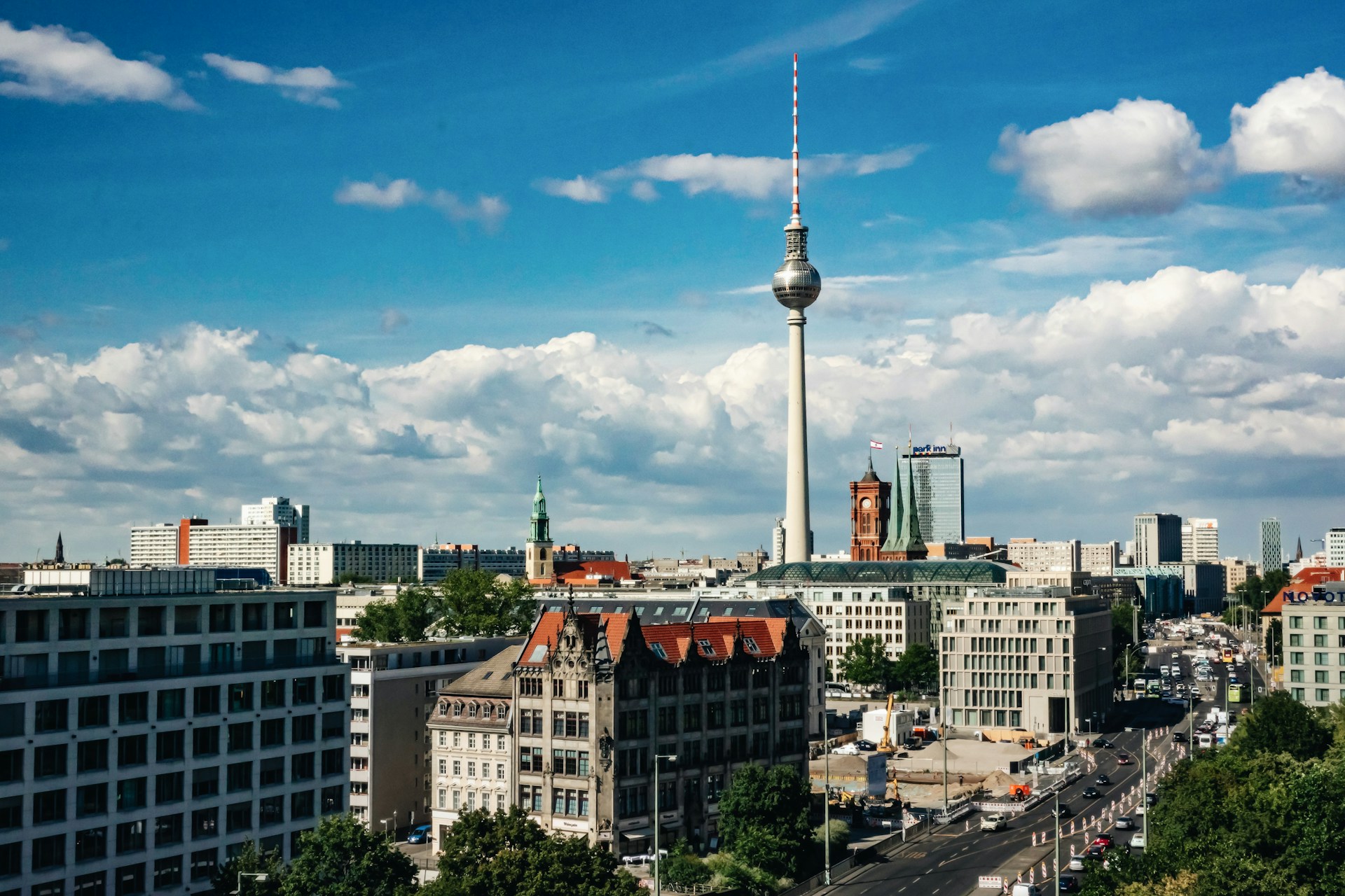 Berlin skyline with the tall Fernsehturm TV tower rising above red-roofed buildings under a blue sky.